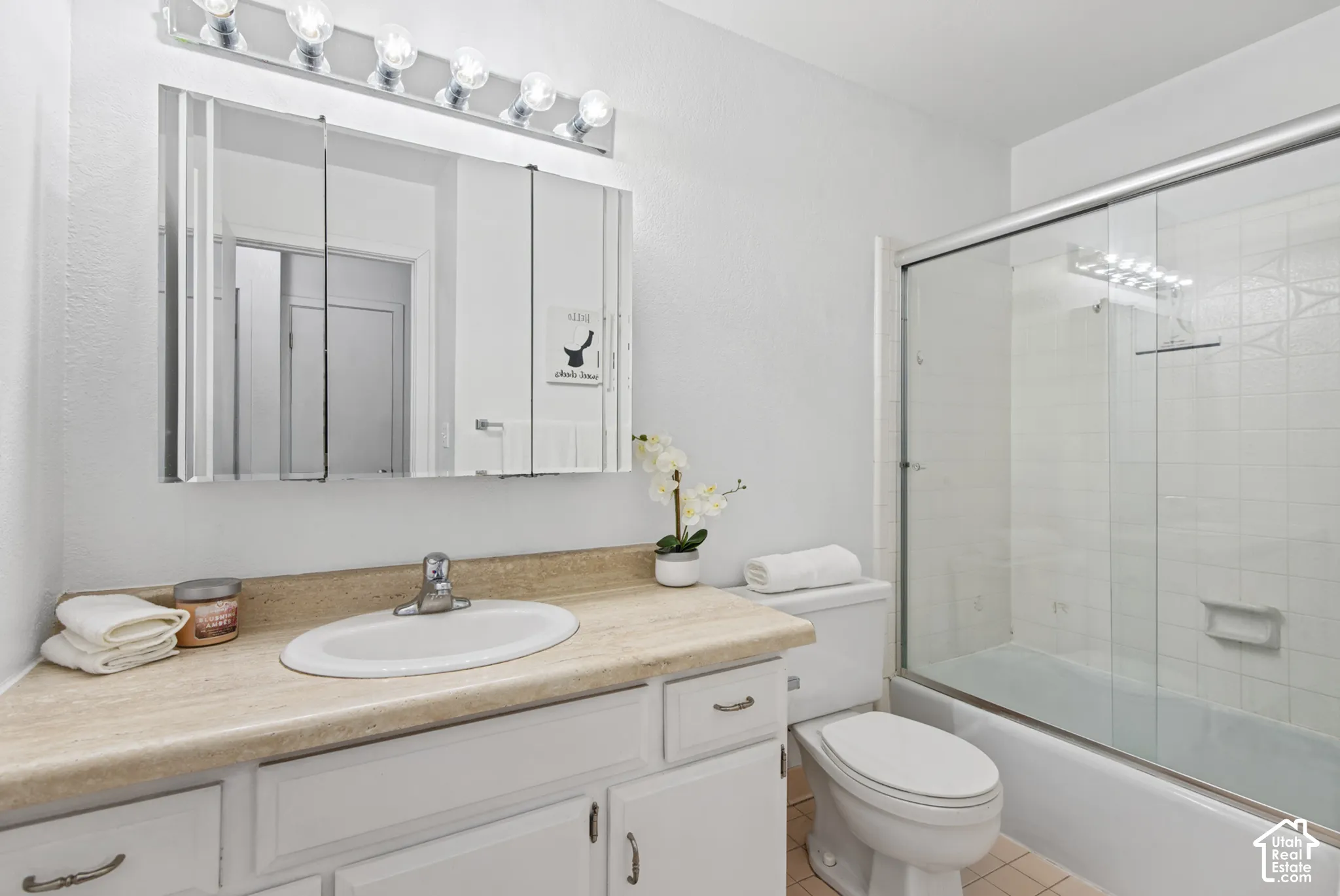 Bathroom featuring a white vanity with a neutral-toned countertop, integrated oval basin, tri-fold mirrored medicine cabinet, six-bulb overhead light fixture, and a glass-enclosed shower and tub combination
