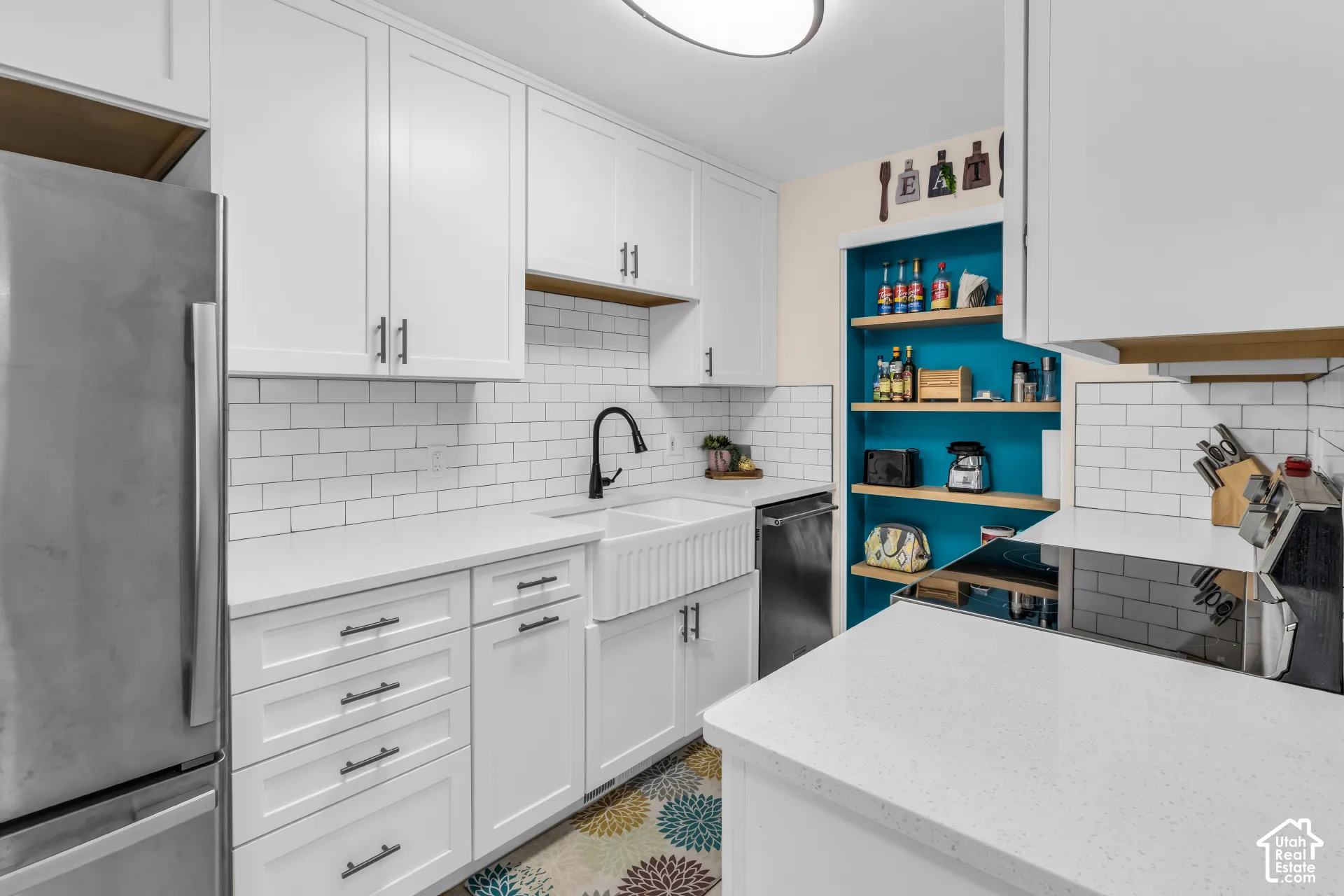 Kitchen featuring white shaker-style cabinetry, white solid-surface countertops, a white farmhouse apron-front sink with a matte black faucet, white subway tile backsplash, and built-in wooden shelving