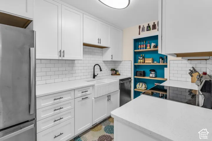 Kitchen featuring white shaker-style cabinetry, white solid-surface countertops, a white farmhouse apron-front sink with a matte black faucet, white subway tile backsplash, and built-in wooden shelving