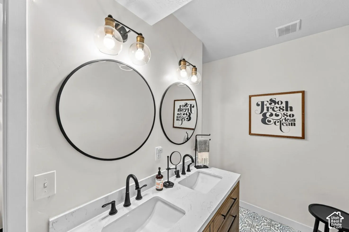 Double vanity featuring a light-toned wood-finish cabinet, quartz countertop with dual undermount sinks, matte black widespread faucets, round mirrors with black frames, and dual globe-style wall sconces
