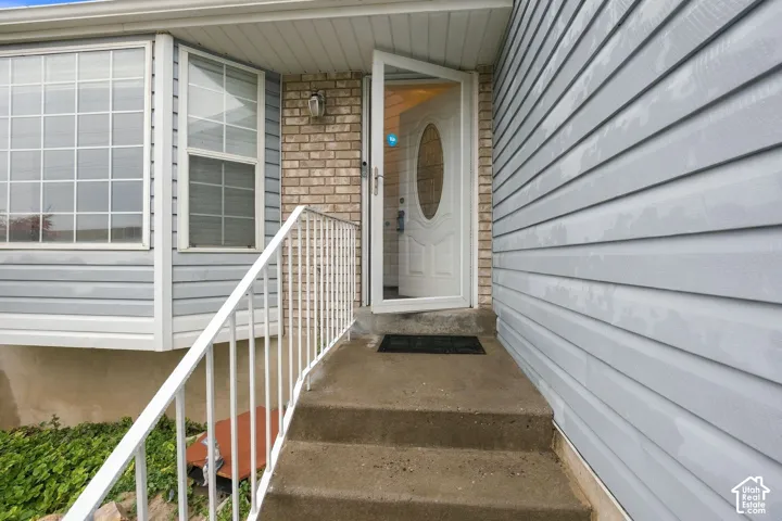 Exterior entrance featuring light blue horizontal siding, tan brick masonry, white metal railing, concrete steps, and a multi-pane bay window