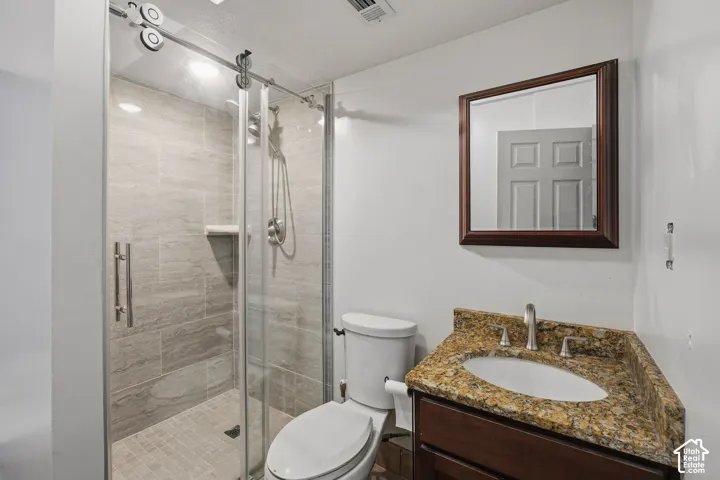Walk-in shower with sliding glass door, stone-tile surround, dark wood-finish vanity, stone countertop with integrated oval sink, and framed wall-mounted mirror