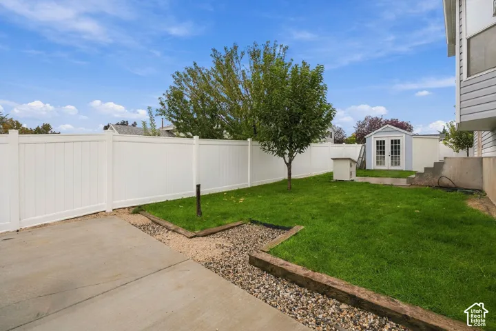 Backyard featuring a concrete patio, white vinyl fencing, timber-bordered landscaping, lawn space, and a detached storage structure