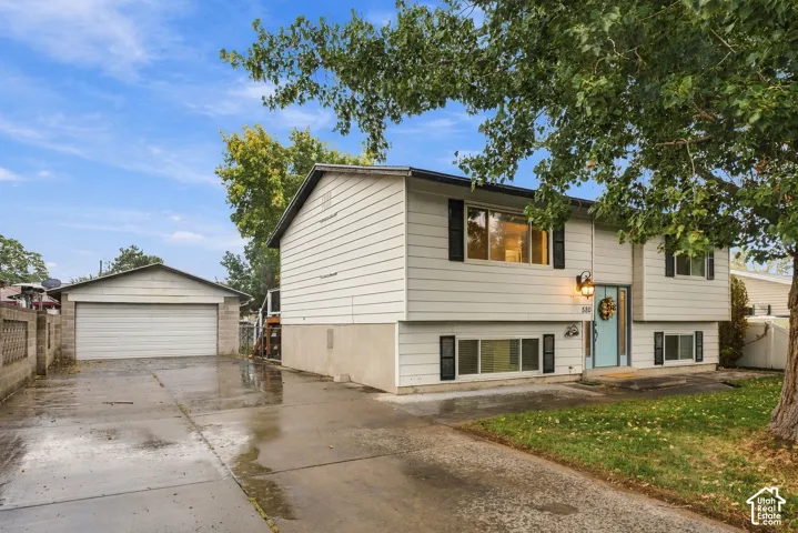 Split-level exterior featuring light-colored horizontal siding, dark decorative shutters, a detached two-car garage, concrete driveway, and mature landscaping