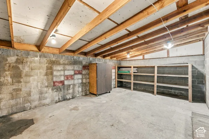 Utility space featuring exposed wood-beam ceiling joists, concrete block walls, concrete flooring, and built-in wood shelving units