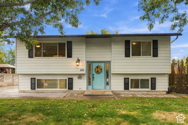 Split-level exterior featuring light-toned horizontal siding, dark decorative shutters, a central entry door with flanking sidelights, and exterior wall-mounted lighting