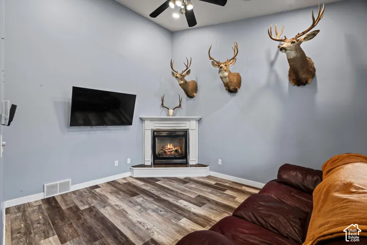 Corner fireplace with a white mantel and stone-finish hearth, accompanied by wood-finish flooring and a ceiling fan with integrated lighting