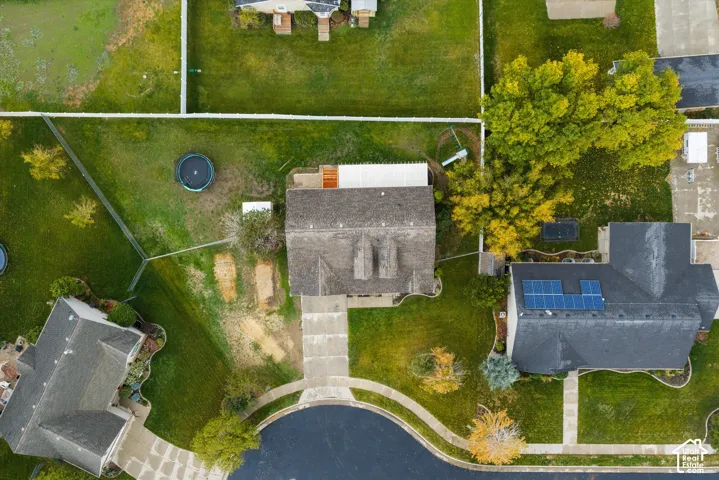 Aerial perspective of a property featuring a concrete driveway, multi-pitched roofline, and expansive fenced lawn with mature landscaping