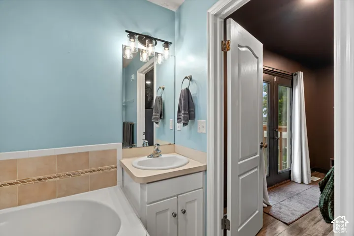 Bathroom featuring a built-in soaking tub with a tiled surround, white vanity cabinetry, wall-mounted light fixture with five glass shades, and a framed mirror