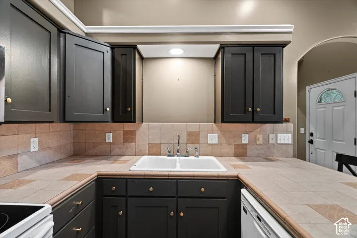 Kitchen area featuring dark-toned cabinetry, a double-basin white sink, tiled countertops with matching backsplash, and recessed lighting