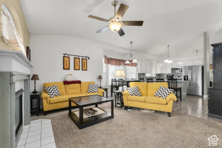 Vaulted ceilings featuring a central paddle fan, gas fireplace with a decorative mantel and tiled hearth, and integrated kitchen cabinetry with stainless steel appliances