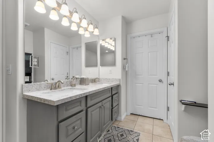 Dual-sink vanity with light-tone stone-finish countertop, cabinetry in a grey finish, large wall-to-wall mirror, multi-bulb overhead lighting, and ceramic tile flooring