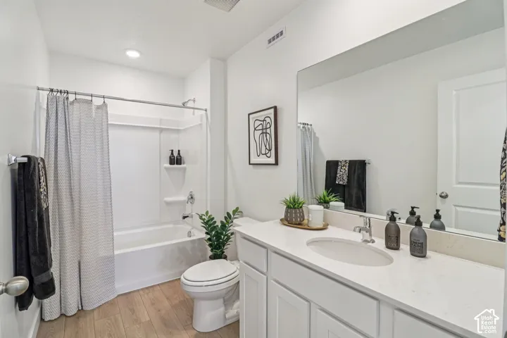 Bathroom featuring a white vanity with a composite countertop, an undermount oval sink, a full-width vanity mirror, a tub-shower combination with integrated shelving, and wood-finish flooring