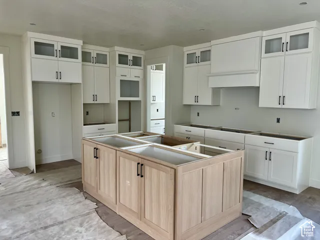 Kitchen featuring white upper cabinetry with glass-front accents, wood-finish base cabinetry, a central kitchen island, and a custom range hood enclosure