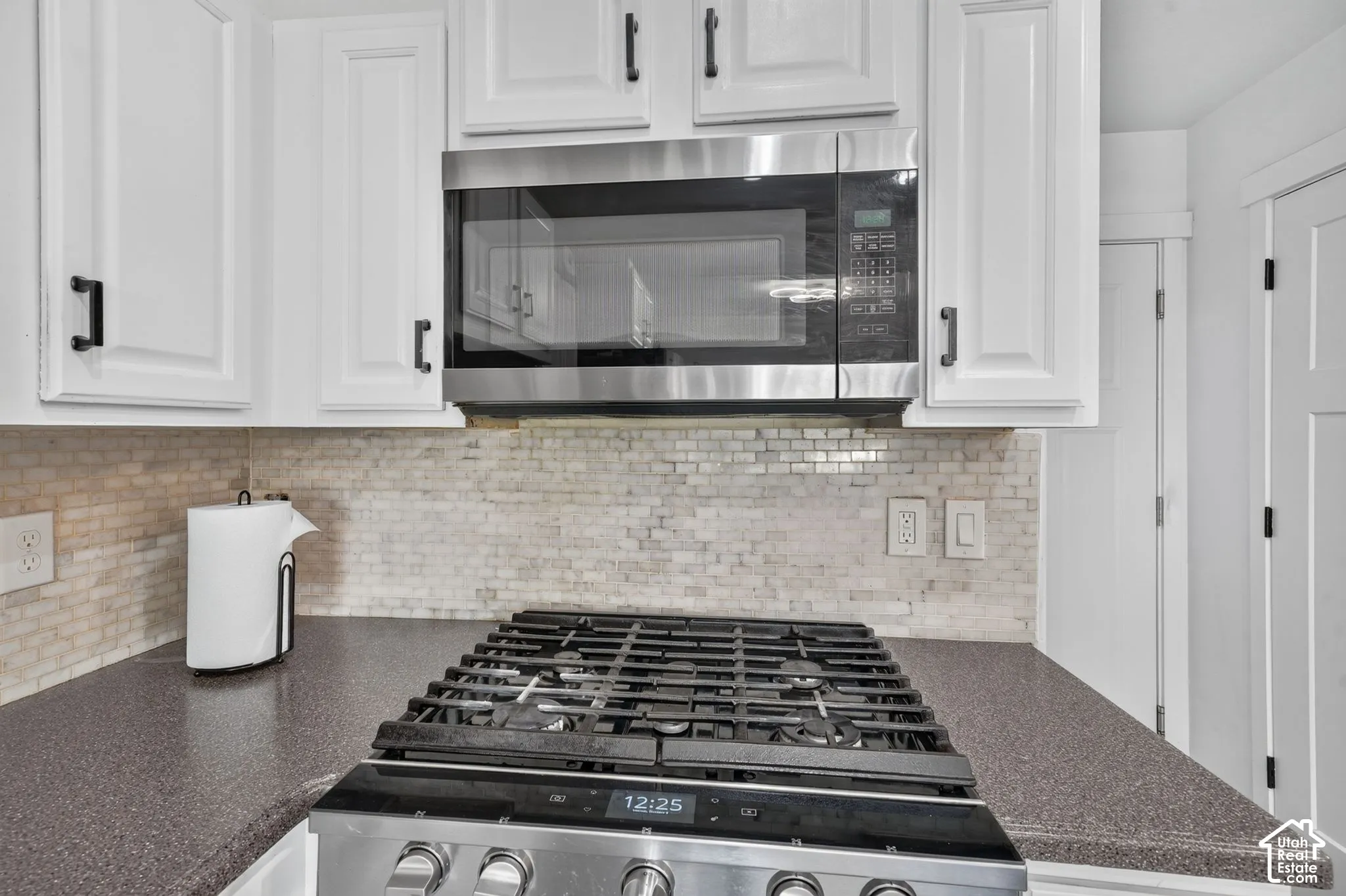 White cabinetry with matte black hardware, stainless steel microwave, stone-tile backsplash, gas range, and solid-surface countertop