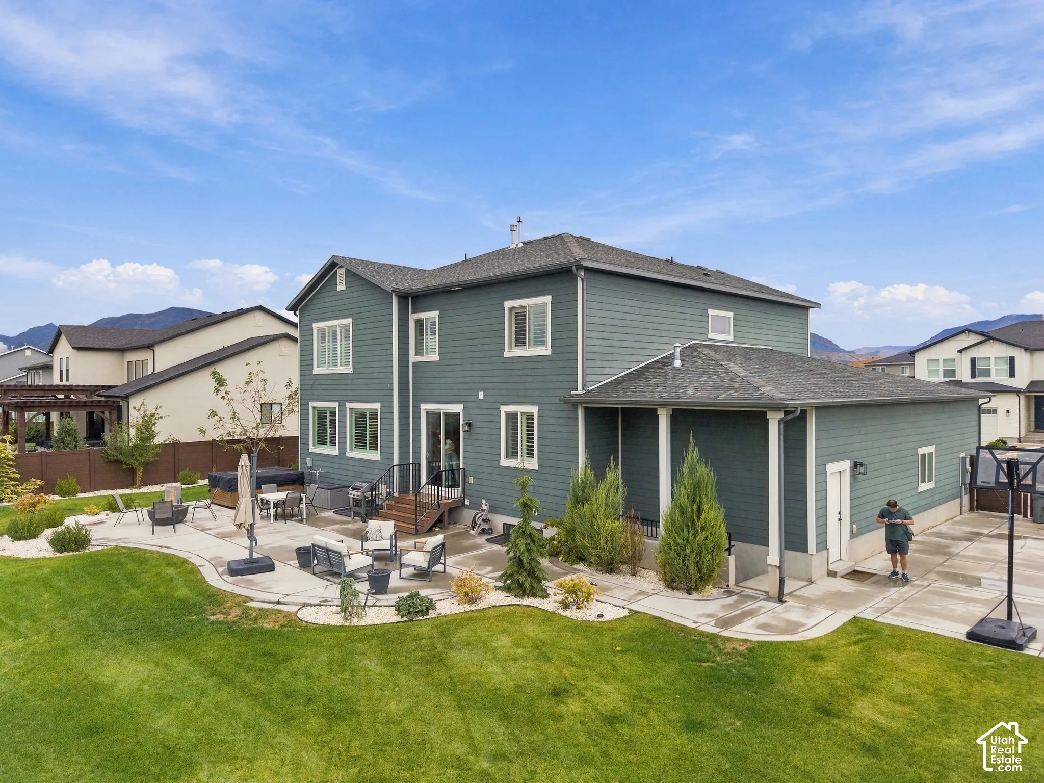 Two-story exterior with horizontal siding, covered side patio, expansive concrete hardscaping, manicured lawn, and mountain backdrop