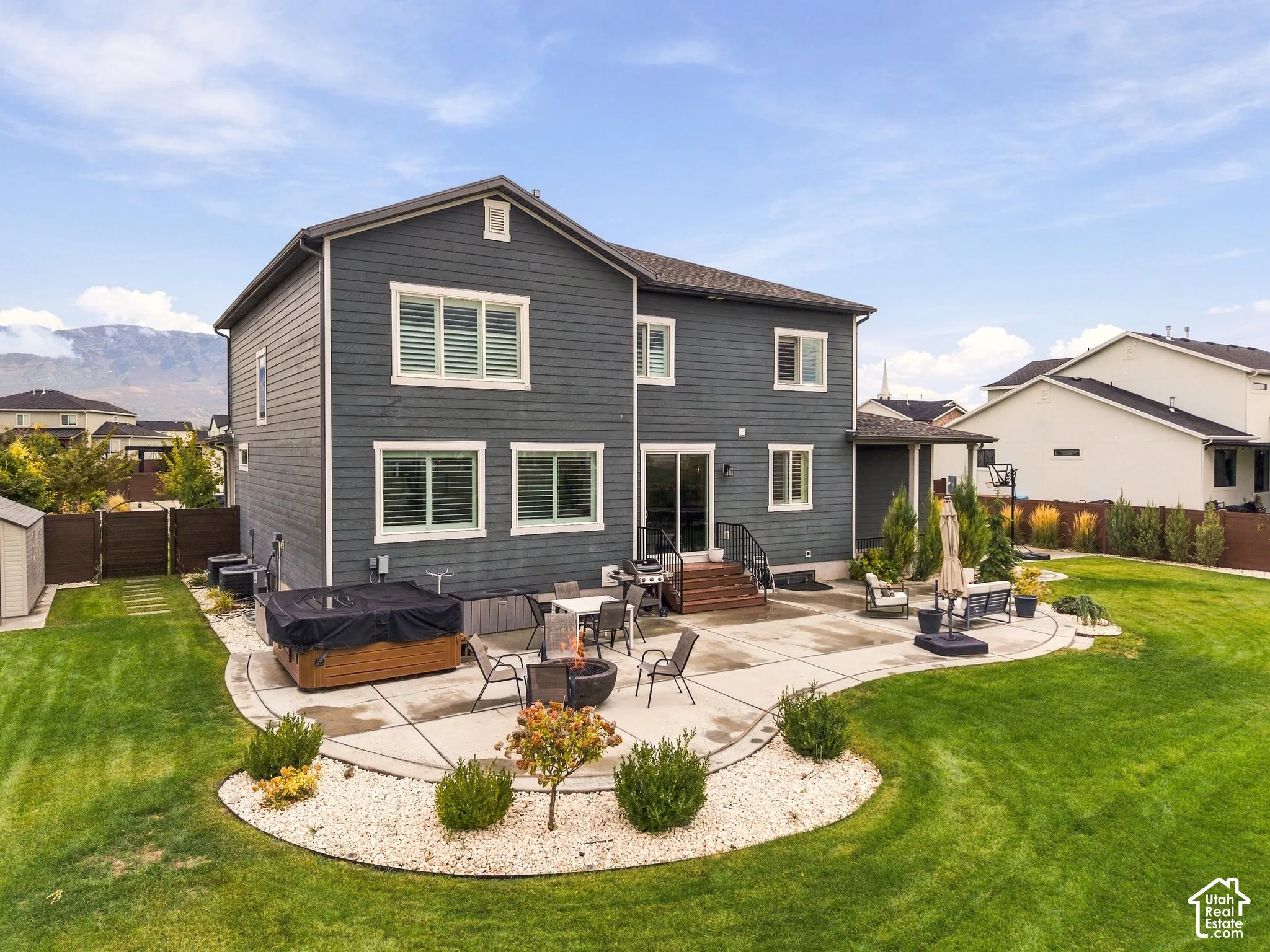 Two-story exterior with horizontal siding, expansive concrete patio, built-in hot tub, landscaping with gravel mulch, and mountain scenery