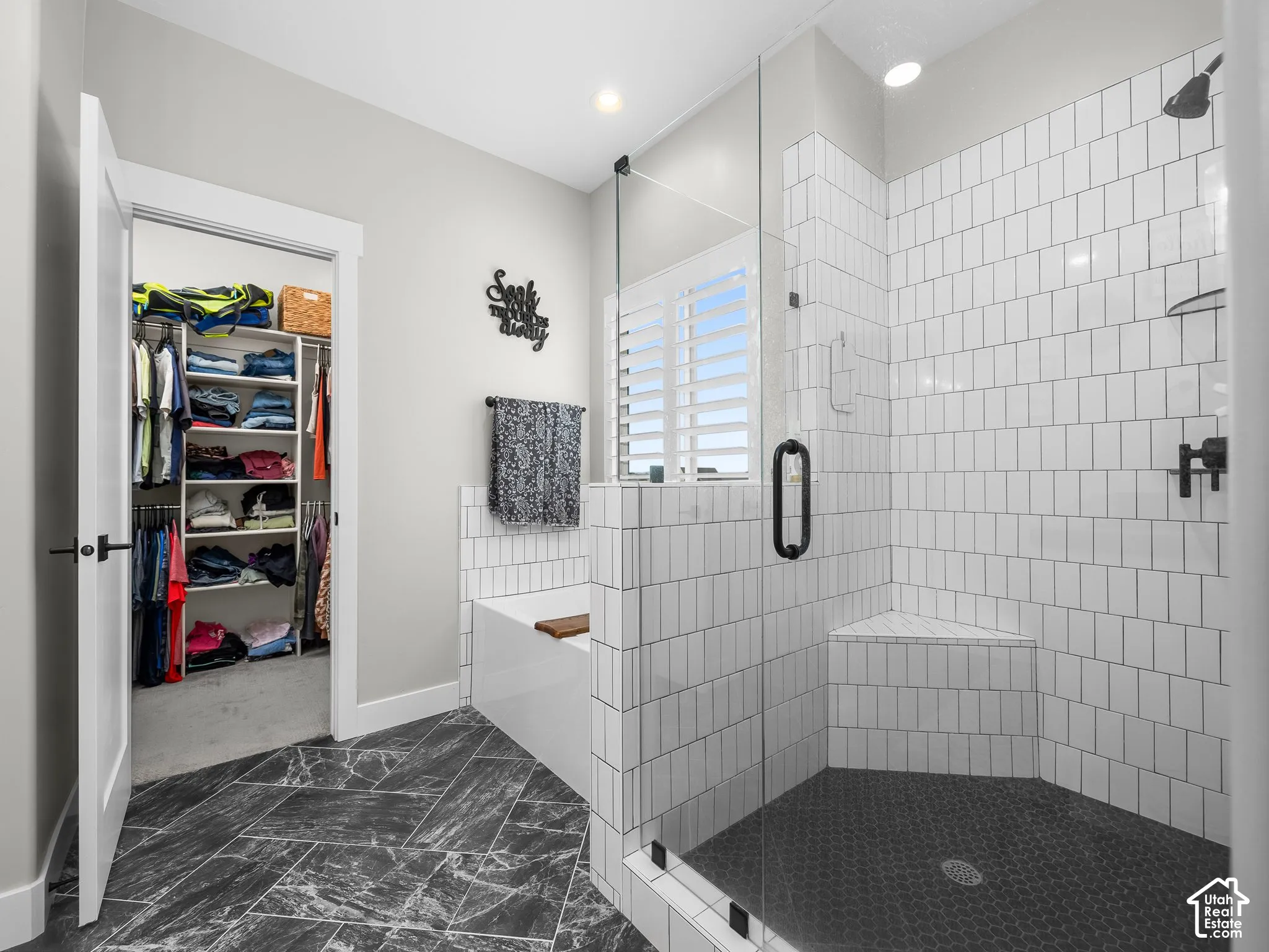 Walk-in shower featuring vertical white subway tile, a built-in bench, and a glass enclosure, complemented by a soaking tub, dark stone-finish tile flooring, and interior plantation shutters