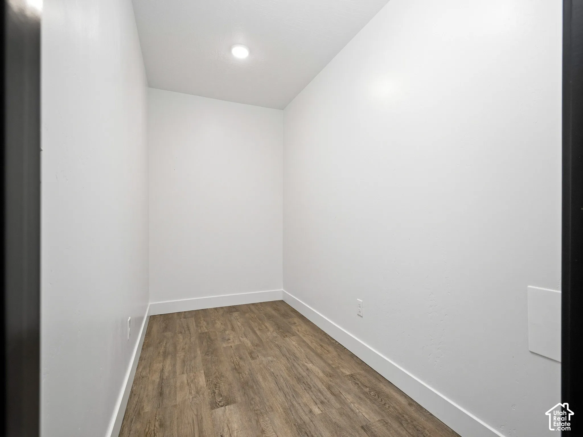 Neutral-toned interior space featuring wood-finish flooring, smooth white walls, and a recessed ceiling light fixture