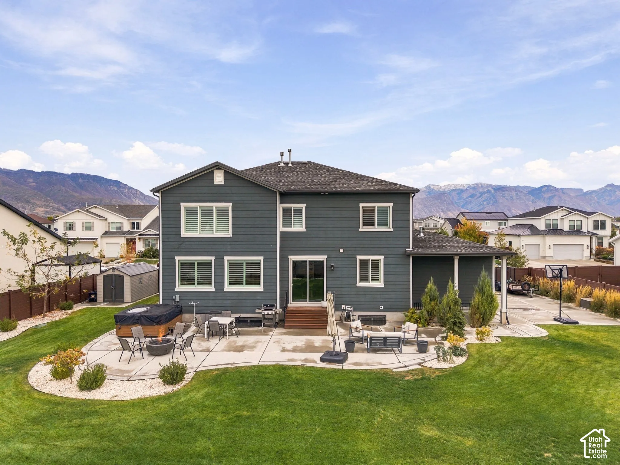 Two-story residence with horizontal siding, expansive concrete patio, built-in hot tub, manicured lawn, and mountain backdrop