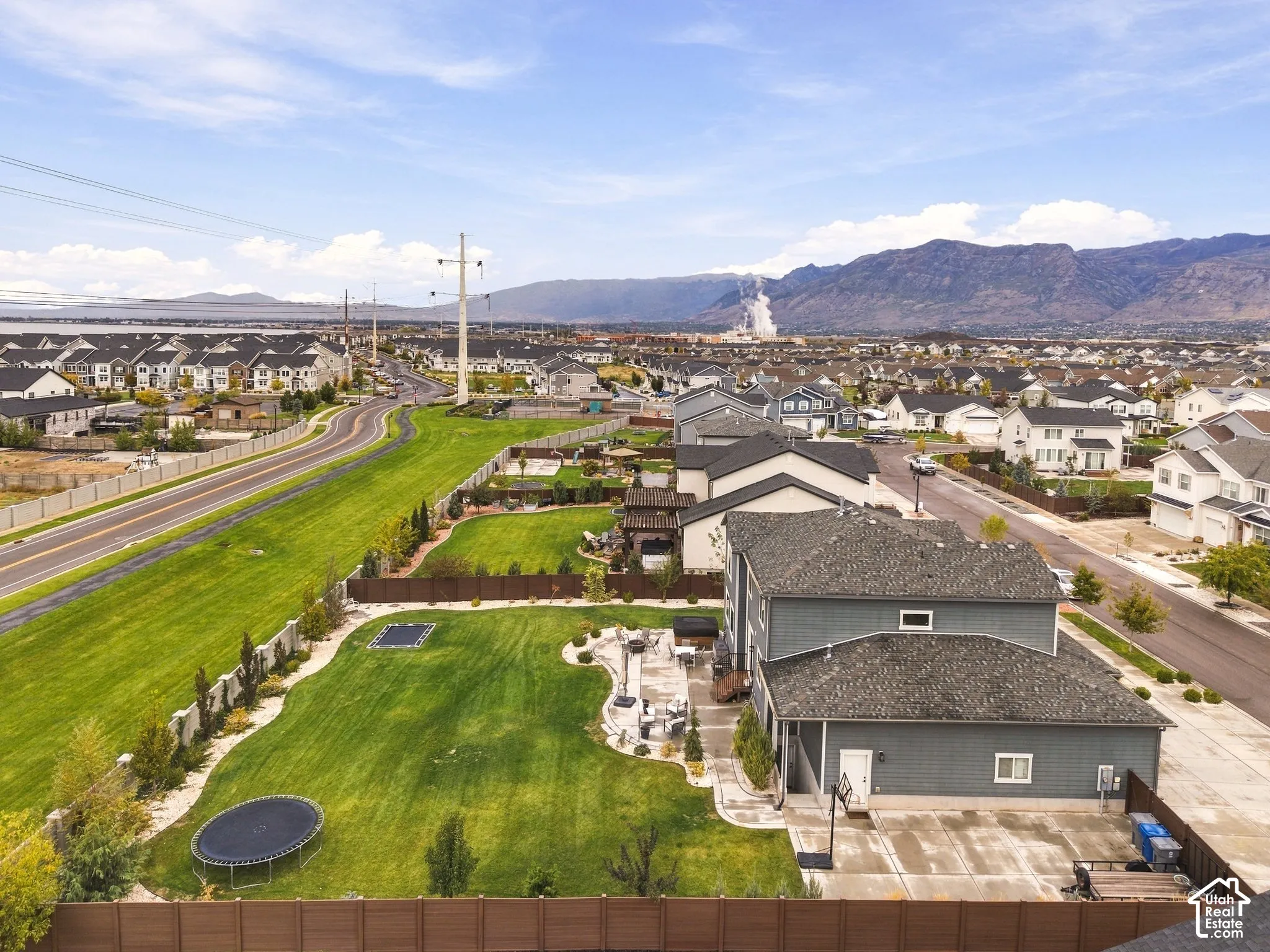 Aerial perspective of a multi-story residence featuring a large manicured lawn, stone-paver patio, privacy fencing, and mountain backdrop