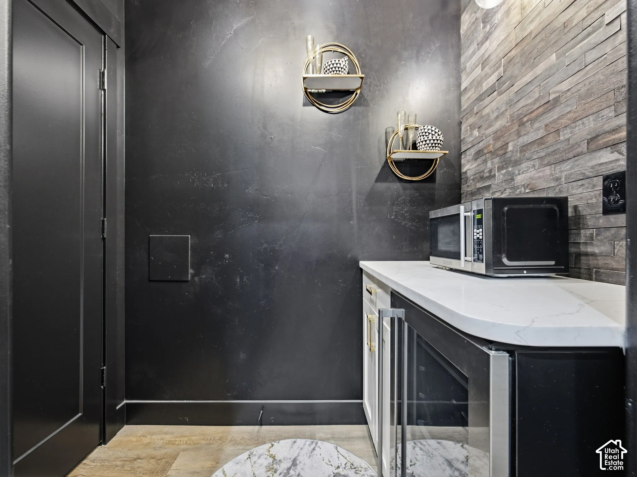 Wet bar featuring a stone-finish accent wall, dark matte wall treatment, white countertop with veining, and a built-in under-counter beverage refrigerator