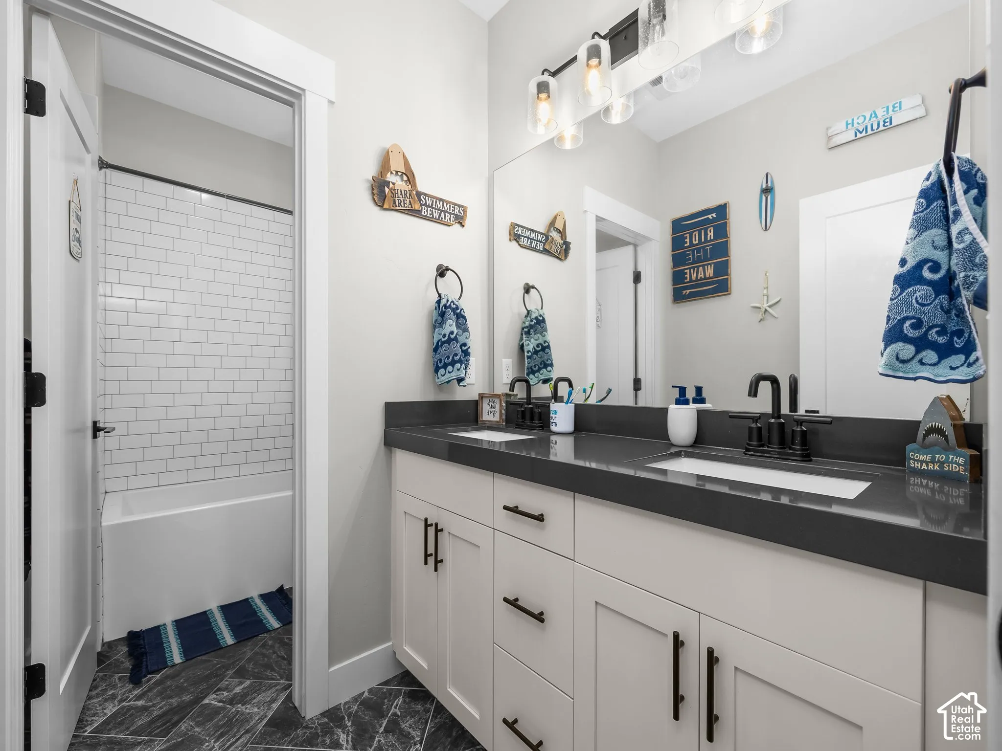 Bathroom featuring a dual-sink vanity with matte black fixtures, quartz countertop, large vanity mirror, four-light glass pendant fixture, and shower-tub combination with white subway tile surround