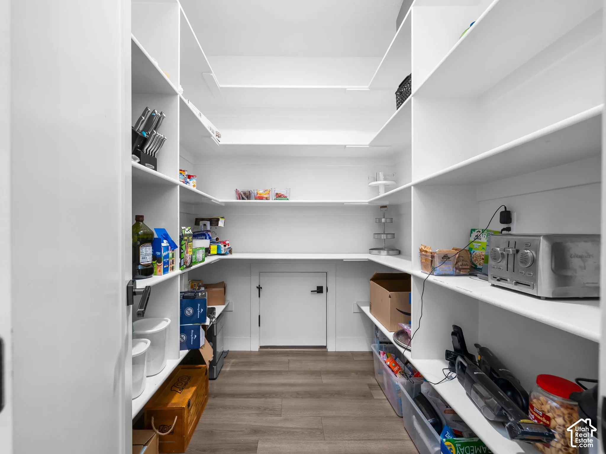Walk-in pantry featuring white wrap-around shelving, wood-finish flooring, and a low-profile service access door with matte black hardware