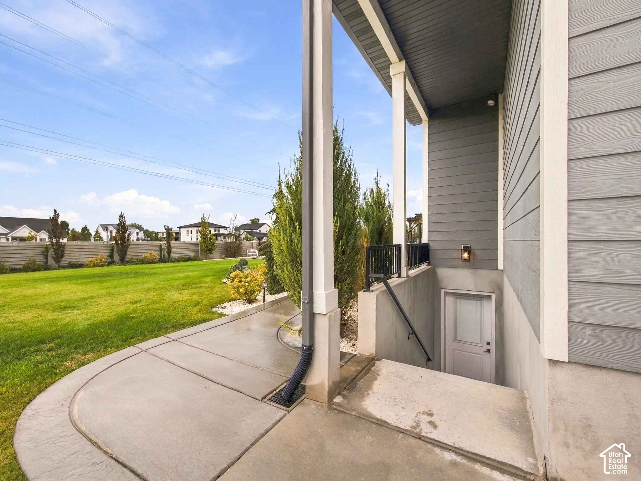 Covered concrete patio with siding exterior, subterranean staircase entrance, metal handrails, and exterior wall-mounted lighting