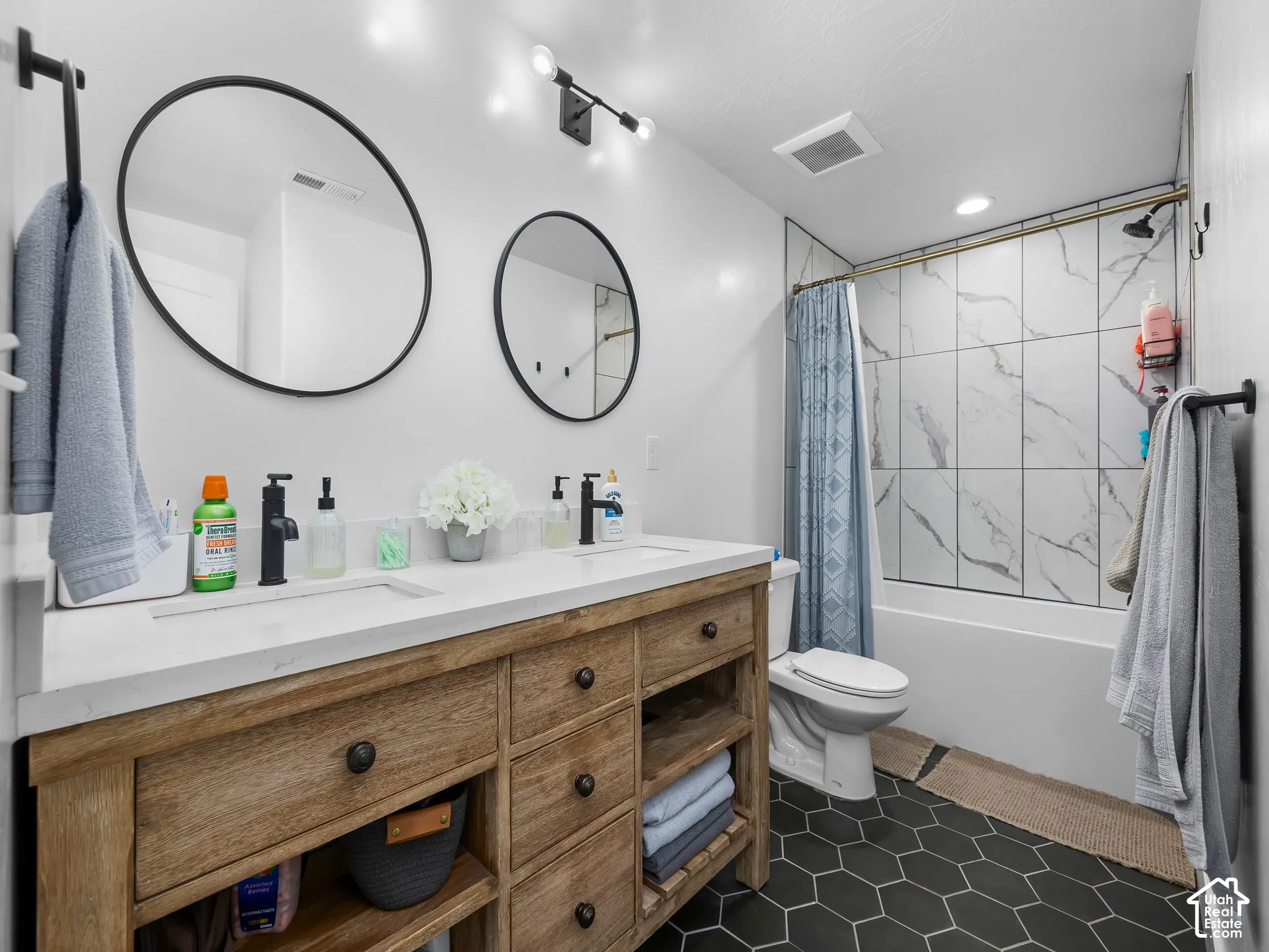 Double vanity featuring a light wood-finish cabinet with integrated shelving, quartz countertop, matte black fixtures, and round mirrors with black frames, complemented by geometric black floor tile and full-height tiled shower surround
