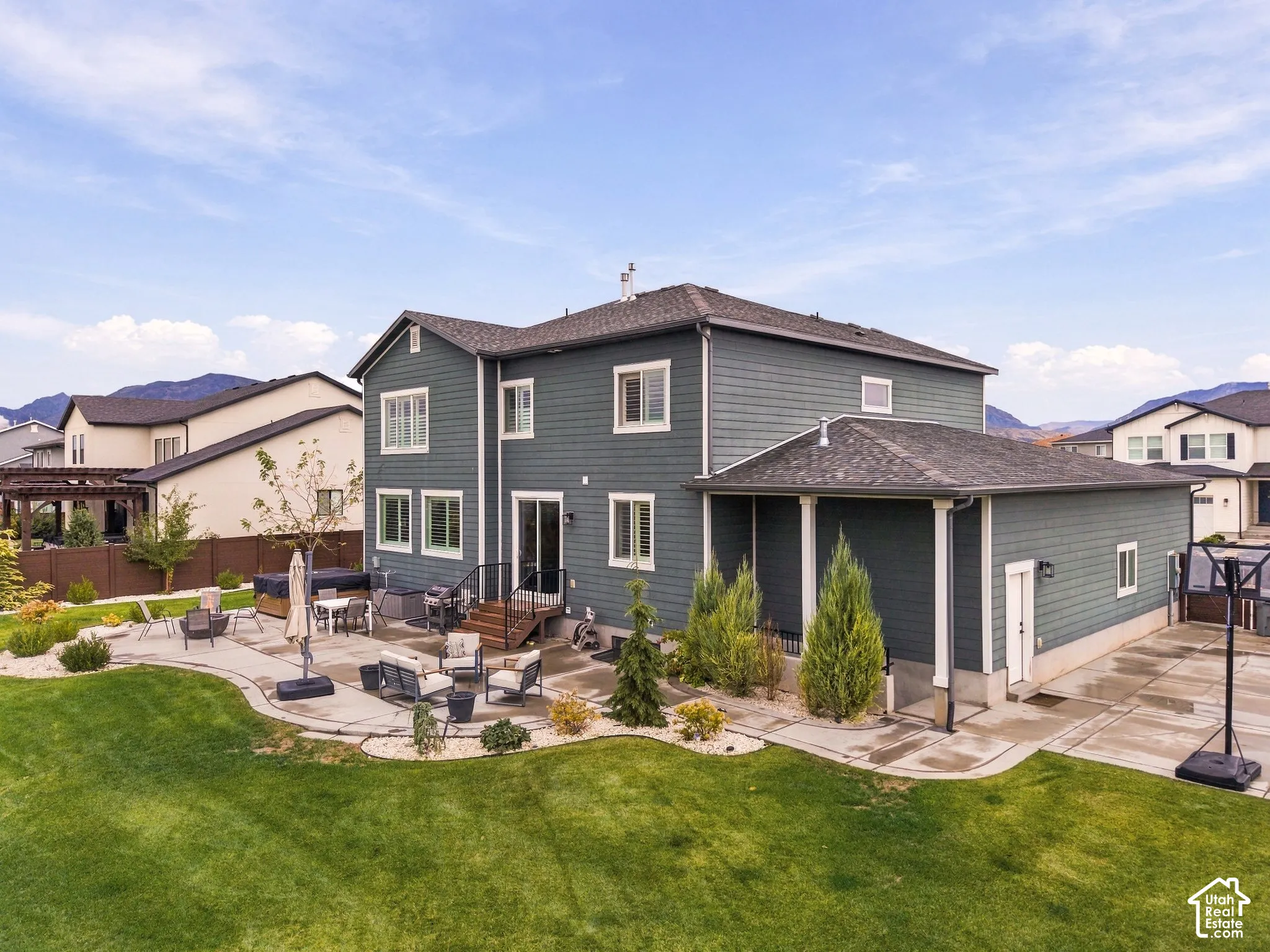 Two-story exterior featuring blue siding, a covered side patio with structural columns, expansive rear stone-paver patio, manicured lawn, and mountain backdrop