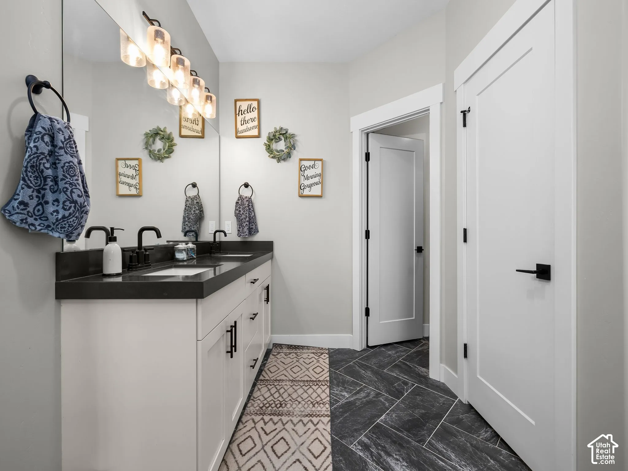 Dual-sink vanity with dark stone countertop, white shaker-style cabinetry, matte black hardware, and herringbone-patterned tile flooring