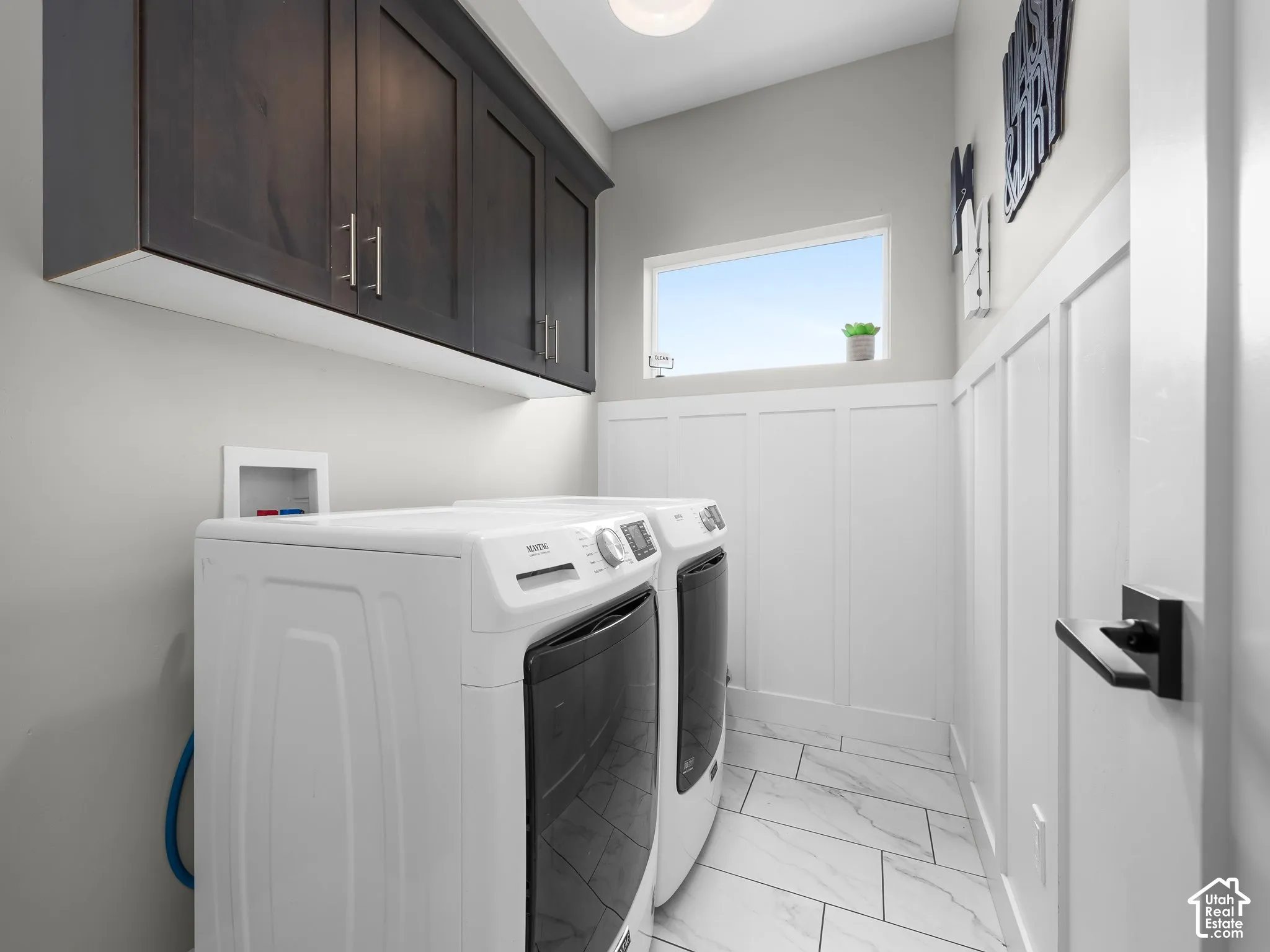 Laundry room featuring dark-finish upper cabinetry, white wainscoting, and stone-finish tile flooring