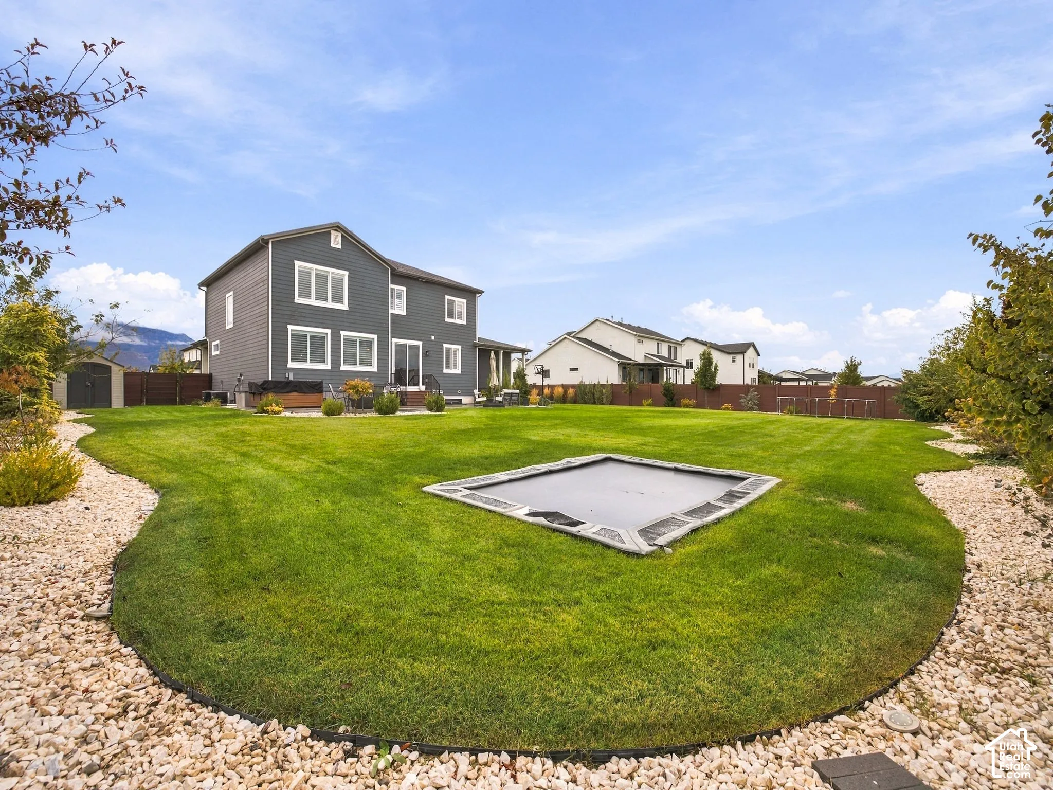 Two-story residence with dark-toned horizontal siding, expansive lawn, stone-accented perimeter landscaping, and full-height privacy fencing