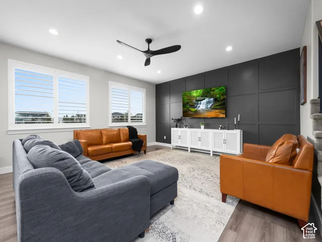 Living area featuring a dark-toned paneled accent wall, wood-finish flooring, recessed lighting, plantation shutters, and a sleek matte black ceiling fan