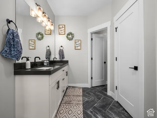 Dual-sink vanity with dark stone countertop, white shaker-style cabinetry, matte black hardware, and herringbone-patterned tile flooring