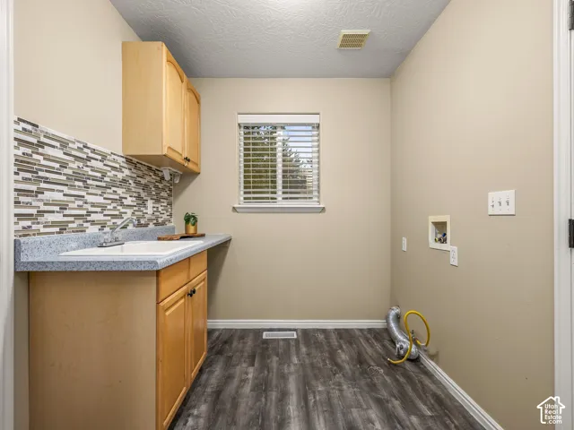 Utility room featuring wood-finish flooring, a neutral-toned tile backsplash, laminate countertop with an integrated basin, upper and lower cabinetry, and window with slatted blinds