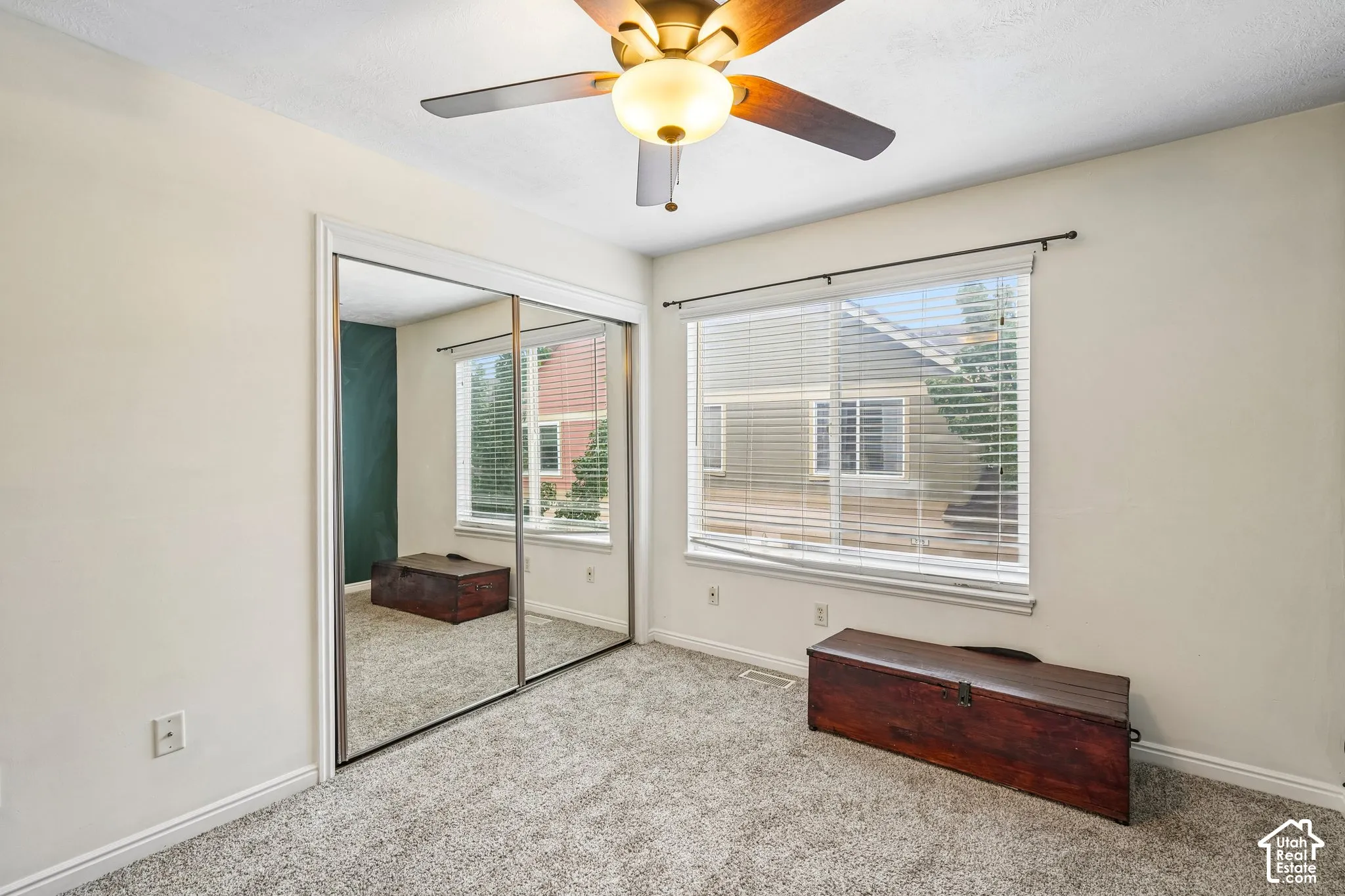 Neutral wall color, carpeted flooring, sliding mirrored closet doors, ceiling fan with integrated lighting, and window with horizontal blinds