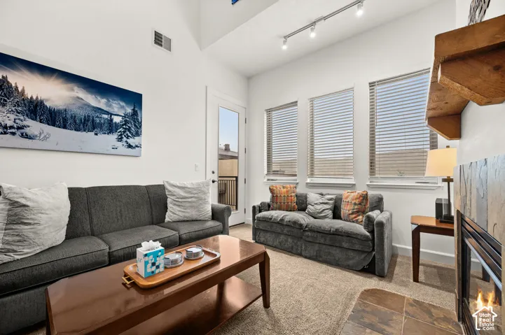 Vaulted ceilings featuring adjustable track lighting, three paneled windows with horizontal blinds, and a fireplace with a stone-surround hearth and natural-finish wood mantel