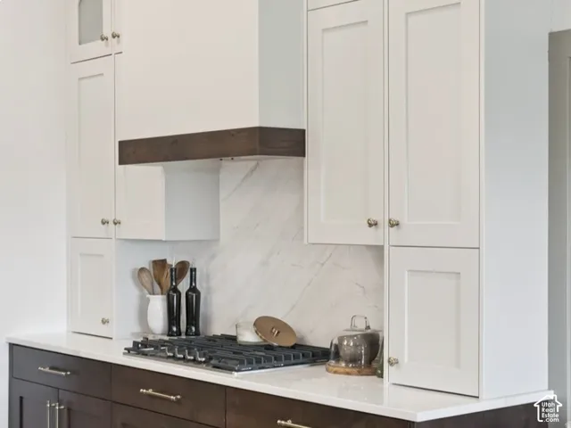 Kitchen featuring white shaker-style upper cabinetry, dark-finish base cabinets, a stone-patterned backsplash, quartz countertops, and a built-in range hood with wood-finish trim