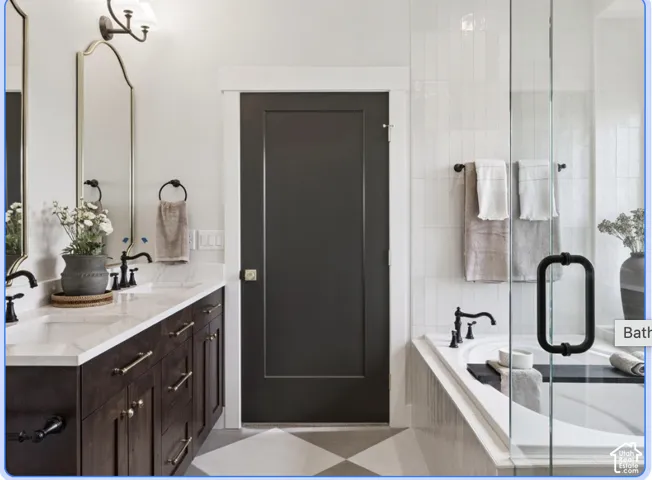 Dual-sink vanity with light-toned countertop, custom dark-finish cabinetry, arched wall mirrors, glass-enclosed soaking tub, and geometric tile flooring