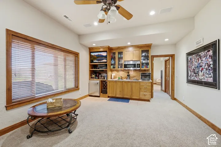Built-in wet bar featuring natural wood cabinetry, stone-finish countertops, stainless steel microwave, and dedicated wine storage, complemented by a ceiling fan and large window with wood-finish trim