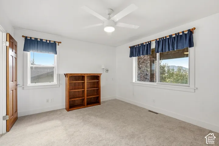 Room interior featuring neutral carpeting, a built-in wooden shelving unit, a white ceiling fan with integrated lighting, and large windows with mountainous scenery