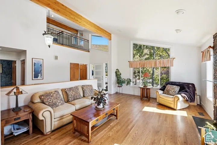 Vaulted ceilings featuring exposed timber beams, expansive windows, wood-finish flooring, a stone-accented fireplace, and an elevated interior balcony with metal railing