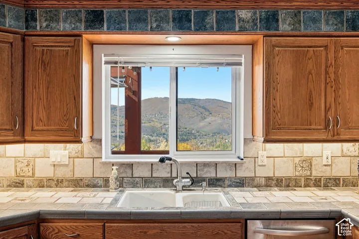 Kitchen featuring custom wood cabinetry, a dual-basin drop-in sink with a gooseneck faucet, a tile countertop, a decorative stone tile backsplash, and a mountain backdrop