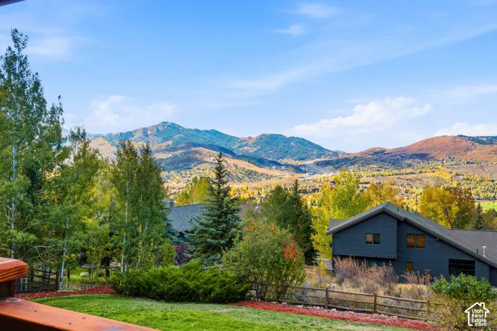 Elevated wooden deck overlooking a natural landscape featuring mature deciduous and coniferous trees, a grass lawn, a split-rail fence, and a backdrop of mountain peaks