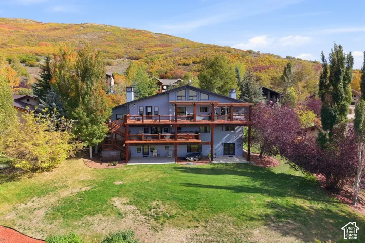 Multi-level home exterior featuring dual stacked wooden decks, a covered ground-level patio, dual stone chimneys, and expansive rear lawn space against a mountain backdrop