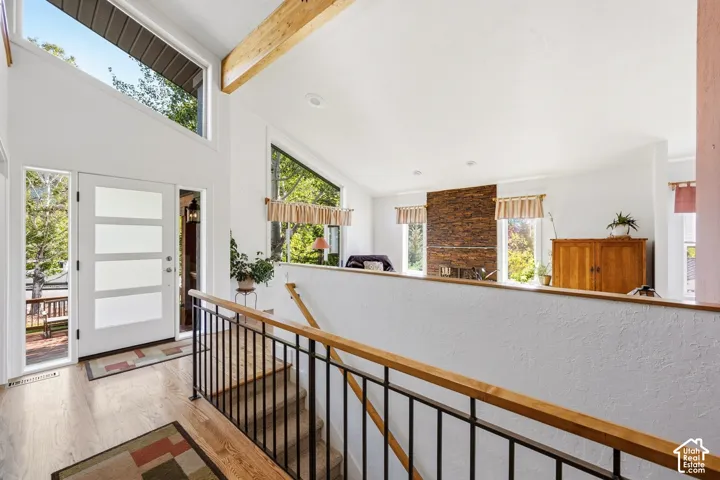 Vaulted ceilings featuring exposed timber beams, expansive windows with clerestory panels, a floor-to-ceiling stone fireplace, and a staircase with a modern metal railing and wood handrail
