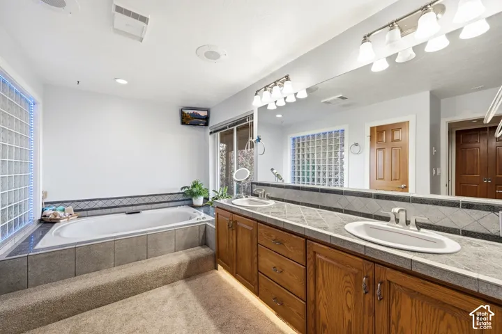 Dual-sink vanity with tile countertop, integrated bathtub, full-width vanity mirror, glass block window detailing, and wood cabinetry