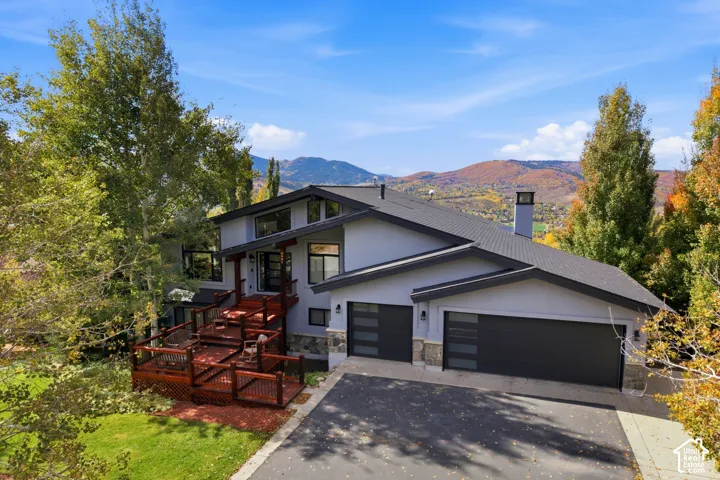 Multi-level exterior featuring a tiered wood deck, stacked stone facade accents, oversized windows, and a three-car garage set against a mountain backdrop